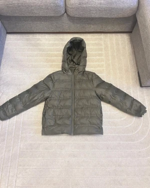 Mörkgrön pufferjacka med huva - Säljer en stilren mörkgrön pufferjacka med huva och dragkedja framtill. Jackan har quiltad design och långa ärmar, perfekt för kalla dagar. Den har en loose passform och är tillverkad i syntetmaterial som håller värmen bra.