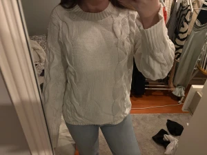 Vit kabelstickad tröja med rund hals - Säljer en vit kabelstickad tröja med rundad hals och långa ärmar. Tröjan har ett klassiskt flätmönster längs fram- och baksida samt ribbade muddar vid hals, ärmslut och nederkant. Perfekt för dig som gillar en clean och cozy stil.