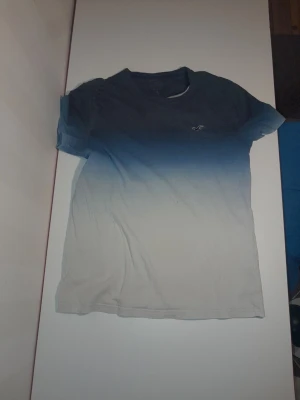 Blåvit t-shirt från Hollister - Snygg t-shirt från Hollister med cool ombre-effekt som går från mörkblå upptill till vit nedtill. Klassisk passform och korta ärmar, med diskret Hollister-logga på bröstet. Perfekt för dig som gillar stilrena och fräscha färgövergångar. Storlek 158
