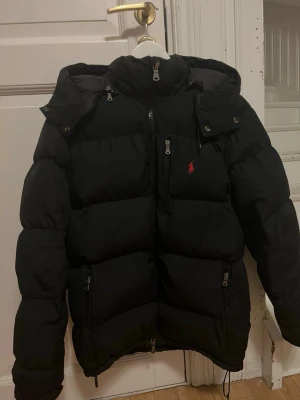 Svart pufferjacka från Polo Ralph Lauren - Säljer en jättesnygg och varm vinterjacka från Polo Ralph Lauren med avtagbar luva. Jackan är köpt på Plick och säljer nu vidare den. Jackan har tagits väl hand om, kontakta mig för mer information/bilder! Ordinarie pris är runt 6000kr