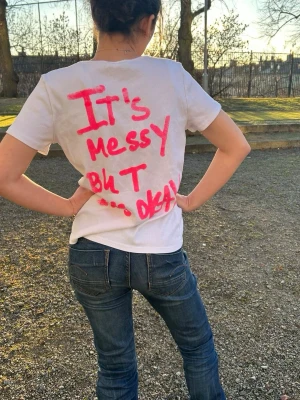 Vit t-shirt med neonrosa screentryck ”Its messy but its okay” - Cool vit t-shirt köpt på sellpy med neonrosa screentryck på ryggen: 'It's messy but it's okay'. T-shirten har korta ärmar och är i ett ribbat material. Perfekt för dig som gillar statement-plagg och vill sticka ut med en unik stil! Rabatt på priset då trycket är lite sämre.