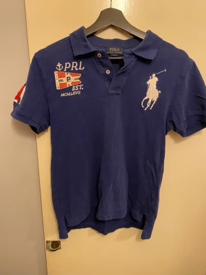 Blå piké från Polo Ralph Lauren - Säljer en blå piké från Polo Ralph Lauren med broderad logga och flaggdetaljer på bröstet och ärmen. Klassisk krage, korta ärmar och knappar framtill. Materialet är mjuk bomull och passformen är normal. Perfekt för dig som gillar sportig och stilren look.