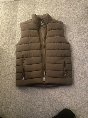 Brun dunväst från Bondelid, S -  ( nypris 1100kr❌, mitt pris 999kr ✅)Snygg brun dunväst från Bondelid i storlek S. Västen har quiltad design med horisontella sömmar, två fickor med dragkedja och hög krage. Perfekt att slänga över en hoodie när det är kyligt ute. Modern och stilren look.
