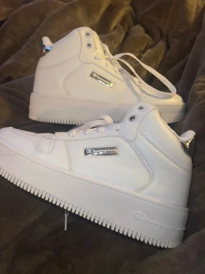 Vita Champion sneakers med platåsula - Säljer ett par vita Champion sneakers i storlek 37.5 med högt skaft och platåsula. Skorna har snörning, silvriga detaljer och Champion-logga på sidan och hälen. Tillverkade i syntetmaterial med en clean och modern look, perfekta för dig som gillar chunky sneakers.