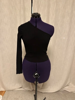 Svart oneshoulder topp från NA-KD - Säljer en svart ribbad topp från NA-KD i oneshoulder-modell med lång ärm på ena sidan. Toppen är tight och har en stilren, asymmetrisk skärning som ger en edgy vibe. Perfekt för dig som vill sticka ut med en cool och modern look.