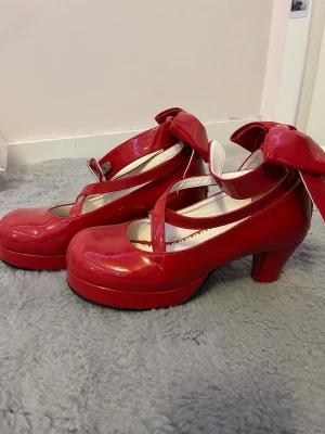 Angelic Imprint Klackar - Supervackra röda klackar med rosett detalj på baksidan från Lolita märket Angelic Imprint! Köpt från second hand men aldrig använt. OBS! har en liten defekt på klacken av förra ägaren (se bild 4) men syns knappast! Dessa klackar är perfekta till er som cosplayar! 