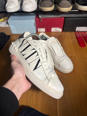 Valentino Garavani skor - Snygga vita Valentino Garavani sneakers i skinn med svart VLTN-logga på sidan och coola nitar runt hälen. Skorna har snörning, rund tå och en silvrig detalj på baksidan. Perfekta för dig som vill sticka ut med exklusiva och trendiga sneakers.