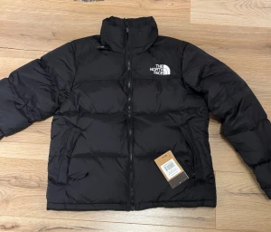 Svart pufferjacka från The North Face - Svart pufferjacka från The North Face med broderad logga i vitt på bröstet och ryggen. Jackan har hög krage, dragkedja framtill och två sidofickor. Materialet är syntet och dun, perfekt för kalla dagar. Klassisk, boxig passform och stilren design.