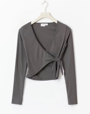 Grå omlottopp från Gina Tricot Young - Snygg grå omlottopp från Gina Tricot med lång ärm och v-ringning. Toppen har knyt-detalj i sidan. Helt oanvänd med lappen kvar💕