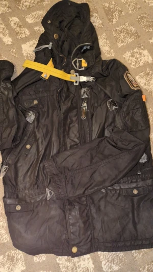 Svart Parajumpers vinterjacka S - Grym svart Parajumpers vinterjacka med ikonisk gul rem och patch på ärmen. Jackan har huva med fuskpäls, flera fickor, dragkedja och knappar. Snygga detaljer och quiltad design gör den både stilren och funktionell. Perfekt för kalla dagar.