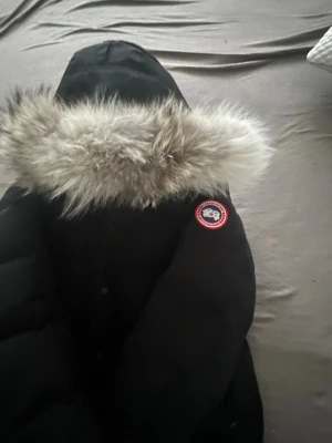 Svart Canada Goose dunjacka med päls - Säljer en svart dunjacka från Canada Goose med ikonisk patch på ärmen och stor huva med fluffig päls. Jackan har en clean look, knappar framtill och är perfekt för kalla dagar. Modellen är klassisk och riktigt varm, perfekt för dig som vill ha både stil och funktion.