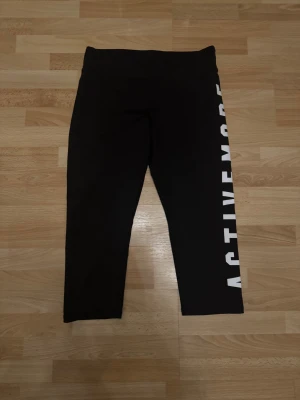 Svarta Capri leggings från H&M Sport - Svarta Capri leggings från H&M Sport med stor vit text 'ACTIVEMODE' längs ena benet. De har en hög midja och är gjorda i stretchigt syntetmaterial som passar perfekt för träning. Snygg och sportig look för dig som gillar att vara aktiv. Storlek 158/164cm, kan även passa xs/s