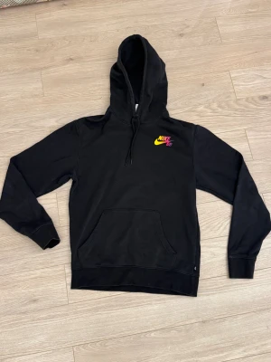 Nike SB hoodie - Svart hoodie från Nike SB i storlek XS med färgstarkt sunset-tryck på ryggen och logga i gult och rosa på bröstet. Tröjan har huva med snörning, magficka och långa ärmar. Perfekt för dig som gillar streetwear och skate-vibe.