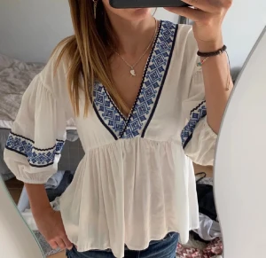 Vit blus med blå broderi och v-ringning - Supersöt vit blus med blått broderat mönster längs v-ringningen och på ärmarna. Blusen har trekvartsärmar med lätt puff och avslappnad passform. Perfekt till jeans eller kjol för en chill och bohemisk vibe.