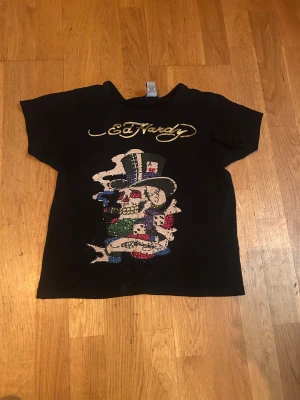 Svart Ed Hardy t-shirt med tryck - Cool svart babytee från Ed Hardy med färgstarkt tryck av döskalle. Guldigt Ed Hardy-logo både fram och bak. Kortärmad och tillverkad i mjuk bomull, perfekt för dig som gillar streetstyle och statement-plagg.