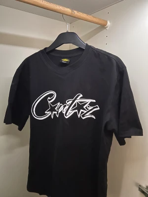 Svart t-shirt med grafiskt tryck - Svart t-shirt från Cortez med stort vitt grafiskt tryck över bröstet i streetstil. Rund halsringning och korta ärmar. Materialet är mjuk bomull som känns skönt mot huden. Perfekt för dig som gillar bold och edgy design.