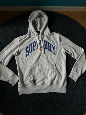 Grå Superdry hoodie med blå tryck - Snygg grå hoodie från Superdry med stor blå logga på bröstet och vita siffror '68' på ena ärmen. Klassisk känguruficka och ribbade muddar. Märke 'Superdry Athletics Dept.' vid fickan. Mjuk bomullsblandning och justerbar huva med snören.