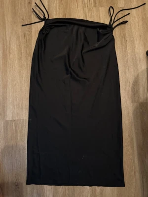 Svart långkjol med snörning - Svart långkjol i mjukt och lätt material med snygga snörningsdetaljer i midjan. Kjolen har en rak och enkel siluett som ger en clean look. Perfekt för dig som gillar minimalistisk stil och vill ha något som är både bekvämt och trendigt.