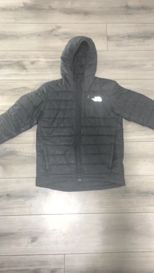 Svart pufferjacka från The North Face storlek 160 - Svart pufferjacka från The North Face i juniorstorlek L. Jackan har huva, dragkedja framtill och ett diskret logotryck på bröstet. Quiltad design och lätt vaddering gör den perfekt för kyliga dagar. Snygg och enkel stil som funkar till streetwear.