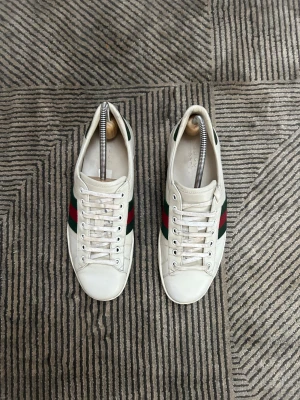 Gucci Ace vita skor - Gucci Ace sneakers i vitt skinn med klassiska gröna och röda ränder på sidorna. Låg modell med snörning och rund tå. Grön detalj i mockaimitation på hälen. Snygga och ikoniska sneakers som lyfter varje outfit.