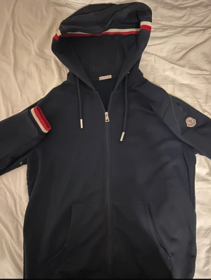 Mörkblå Moncler hoodie med dragkedja - Snygg mörkblå hoodie från Moncler med hel dragkedja, huva och snörning med metalländar. Tröjan har långa ärmar med ribbade muddar i rött, vitt och blått samt Moncler-logga på ärmen. Perfekt för dig som gillar exklusiva streetwear-vibbar.