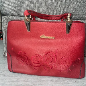Röd handväska med broderier  - Snygg röd handväska med guldfärgade detaljer och broderade rosor på framsidan. Väskan har två handtag, dragkedja baktill och är tillverkad i skinnimitation. Den har även 4 innerfickor.