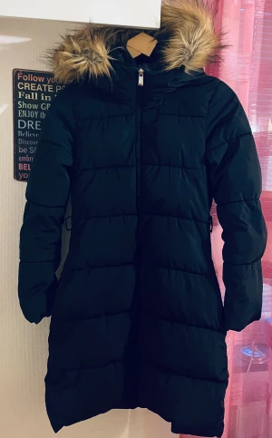 Svart lång jacka  från Lindex - Säljer en svart lång jacka från Lindex i storlek XS. Jackan har huva med fluffig fuskpäls, dragkedja framtill och quiltad design. Perfekt för kalla dagar med sin varma och stilrena look. Passformen är normal och ärmarna är långa.