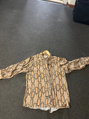 Retro mönstrad skjorta från MD' trend - Snygg retroinspirerad skjorta från MD' trend i storlek XXL. Skjortan har ett unikt geometriskt mönster i beige, svart och vitt, långa ärmar och knäppning framtill. Materialet är glansigt och känns lyxigt mot huden.