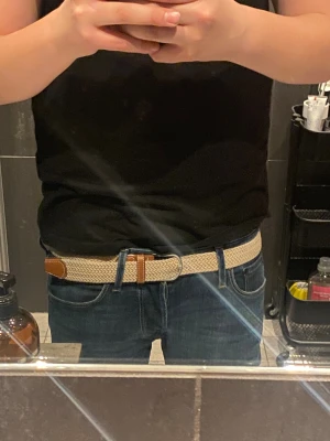 Beige flätat bälte med läderdetaljer - Snyggt beige flätat bälte med bruna läderdetaljer och silvrigt spänne. Bältet är elastiskt och har en stilren look som passar till jeans eller chinos. Perfekt accessoar för att lyfta din outfit med en clean och modern touch.