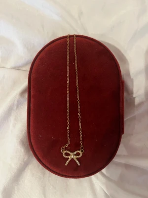 Guldfärgat halsband med rosett och stenar - Supersött halsband i guldfärg med en liten rosett som hänge, täckt av gnistrande stenar. Kedjan är tunn och elegant, perfekt för att ge en extra touch till din outfit. Rosetten ger en gullig och trendig vibe.