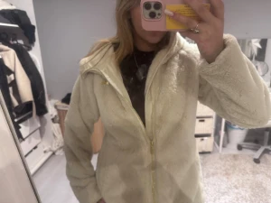 Beige teddyjacka  - Supermjuk och fluffig beige teddyjacka med huva och guldig dragkedja. Jackan har en avslappnad passform och är perfekt för kyliga dagar. Fodrad insida och diskret guldmärke på bröstet. Snygg och enkel att matcha till olika outfits.