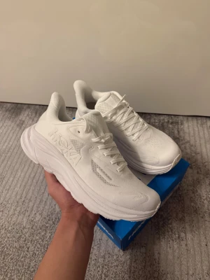 Hoka vita sneakers med tjock sula - Säljer ett par fräscha vita Hoka sneakers med chunky platt sula och snyggt broderad logga på sidan. Skorna har ventilerande meshpaneler, snörning och rund tå. Perfekta för dig som gillar stilrena och sportiga skor med extra komfort.