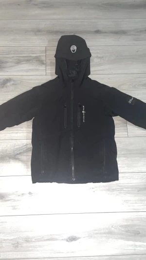 Svart dunjacka Sail Racing med huva - Svart dunjacka från Sail Racing med huva och flera dragkedjor framtill. Jackan är quiltad på insidan och har tekniska egenskaper som vattentät, vindtät och andas. Primaloft-foder och återvunnet material. Snygg logga på huvan och bröstet.