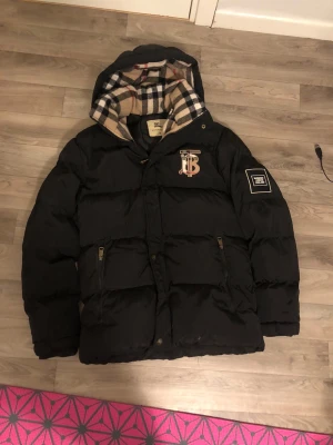 Svart pufferjacka från Burberry  - Svart pufferjacka från Burberry med ikonisk rutig huva i beige, svart och rött. Jackan har dragkedja, två fickor med zip, Burberry-logga på bröstet och patch på ärmen. Perfekt för kalla dagar med sin fluffiga och stilrena look. Jag tar gärna emot betalningen ifrån vinted