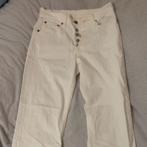 Levi's 501 vita jeans - Säljer ett par klassiska Levi's 501 jeans i vit färg. De har raka ben, knappgylf och fem fickor med snygga nitar. Jeansen är tillverkade i bomull och har den ikoniska Levi's-lappen bak samt röd Levi's-tag vid bakfickan. Aldrig använda.