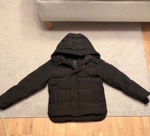 Svart pufferjacka från Canada Goose - Svart pufferjacka från Canada Goose med stor huva, diskret logotyp på ärmen och dragkedja framtill. Jackan har quiltad design, två stora fickor och är långärmad. Perfekt för kalla dagar och har en clean, modern look.