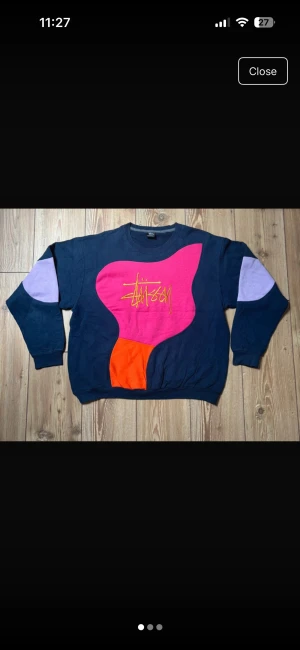 Rework Stüssy färgglad crewneck tröja - Unik crewneck från Stüssy med mörkblå bas och stora färgblock i rosa, orange och lila. Stüssy-logga broderad i gult på bröstet. Tröjan har långärmade ärmar med lila detaljer på armbågarna och är gjord i mjuk bomull. Perfekt statement-plagg för dig som gillar streetwear.🥰