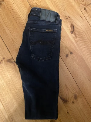 Mörkblå Nudie jeans W28 L30 - Snygga mörkblå jeans från Nudie i klassisk femficksmodell. Jeansen är tillverkade av 100% ekologisk bomull och har en slim passform med subtila detaljer på bakfickan. Perfekt för dig som gillar stilrena och tidlösa jeans.