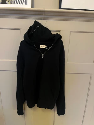 Svart hoodie med half zip från East West - Svart hoodie från East West i storlek M. Tröjan har en stor huva och en snygg half zip-dragkedja framtill. Tillverkad i mjuk bomull, perfekt för chill dagar. Långärmad och med klassisk loose passform.