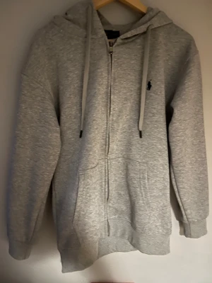 Grå zip-hoodie från Polo Ralph Lauren - Snygg grå hoodie med dragkedja från Polo Ralph Lauren. Klassisk design med svart broderad logga på bröstet, huva med dragsko och stora fickor framtill. Mjuk bomullsblandning som är skön att ha på sig. Perfekt för en chill och stilren look.
