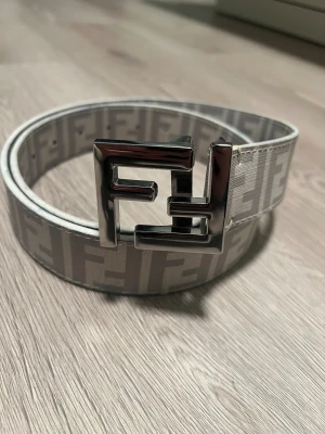 Fendi vit FF-logo bälte 110 cm - Säljer ett snyggt Fendi-bälte i vitt med det klassiska FF-monogrammet i ljusgrått. Bältet har en stor, blank metallspänne i silver med Fendi-logga och texten 'Made in Italy'. Materialet är präglat läder med exklusiv känsla. Använt 2 gånger och det finns ingen skada på bältet. 