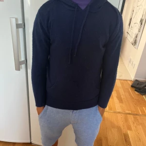 Mörkblå hoodie i kashmir från Maison Cashmere storlek XS - Snygg mörkblå hoodie från Maison Cashmere i mjuk kashmir. Tröjan har huva med snörning, klassisk kanguruficka och långärmad passform. Perfekt för dig som gillar stilren och chill look med lyxig känsla. Passar dig som vill ha något extra mjukt och varmt. Har en liten defekt som ni kan se men inget märkvärdigt eller som speciellt syns