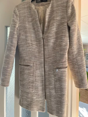 Beige kavajjacka från ZARA - Snygg beige kavajjacka från ZARA med diskret rutigt mönster och rak passform. Jackan har dragkedja framtill, två fickor med dragkedja och är helfodrad. Materialet är strukturerat och ger en exklusiv känsla. Perfekt för dig som gillar stilrena och moderna plagg.