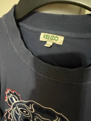 Mörkblå sweatshirt med tiger från Kenzo - Snygg mörkblå sweatshirt från Kenzo med ikonisk broderad tiger på bröstet och röd Kenzo-text. Tröjan har rund hals, ribbade muddar och lång ärm. Perfekt för dig som vill sticka ut med ett coolt statement-plagg. 