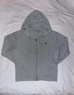 Grå zip hoodie från Polo Ralph Lauren - Snygg gråmelerad hoodie med dragkedja från Polo Ralph Lauren. Klassisk huva med dragsko, två fickor fram och ikonisk mörkblå broderad logga på bröstet. Mjuk bomullsblandning som är skön att ha på sig och passar perfekt till jeans eller joggers.