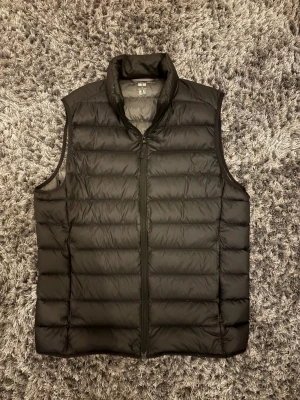 Svart dunväst från Uniqlo, storlek S - Svart, quiltad dunväst från Uniqlo Ultra Light Down. Västen har hög krage, dragkedja framtill och en clean, minimalistisk look. Perfekt för lager-på-lager och enkel att bära tack vare sin lätta känsla.