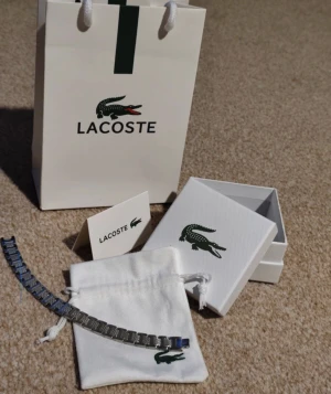 Lacoste silverfärgat armband - Stilrent armband från Lacoste i silverfärgad metall med rektangulära länkar och diskret textur på vissa partier. Kommer med originalpåse, ask och presentpåse med klassiska Lacoste-loggan. Oanvänd ligger i plast, ett till ett, fraktar snabbt .Perfekt accessoar för att lyfta din outfit.