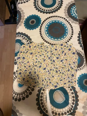 Blommig gul korsett-topp med volang - Supersöt korsett-topp med smala axelband och blommigt mönster i blått, lila och rosa på gul botten. Toppen har markerade kupor och volangdetalj nedtill, perfekt för en somrig look. Materialet känns lätt och luftigt, troligen bomull eller polyester. Finns ett band/snöre som jag ej vet det är till men det löser man! 