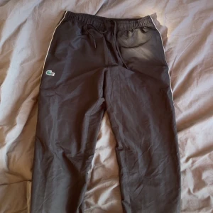 Lacoste byxor - Svarta byxor från Lacoste med vit piping längs sidorna och klassisk krokodillogga på vänster ben. Byxorna har elastisk midja med snörning och är tillverkade i ett mjukt, syntetiskt material. Perfekta för en chill och sportig look.ny pris 1600kr