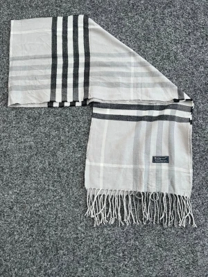 Rutig ljusgrå halsduk från Burberry - Riktigt schysst halsduk från Burberry i ljusgrått med klassiskt rutigt mönster och svarta detaljer. Gjord i 100% lammull, mjuk och skön med fransar i ändarna. Perfekt för att höja din outfit 